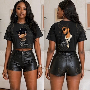 May include: A black cropped t-shirt featuring a graphic print of a person wearing a baseball cap, sunglasses, and a chain necklace, paired with black leather-like shorts. The shirt displays a signature graphic below the portrait.
