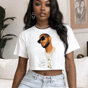 May include: A white short-sleeved crop top featuring a graphic print of a man wearing black sunglasses and multiple layered gold chains. Below the graphic is a black signature. The shirt has a crew neckline and a cropped hemline.