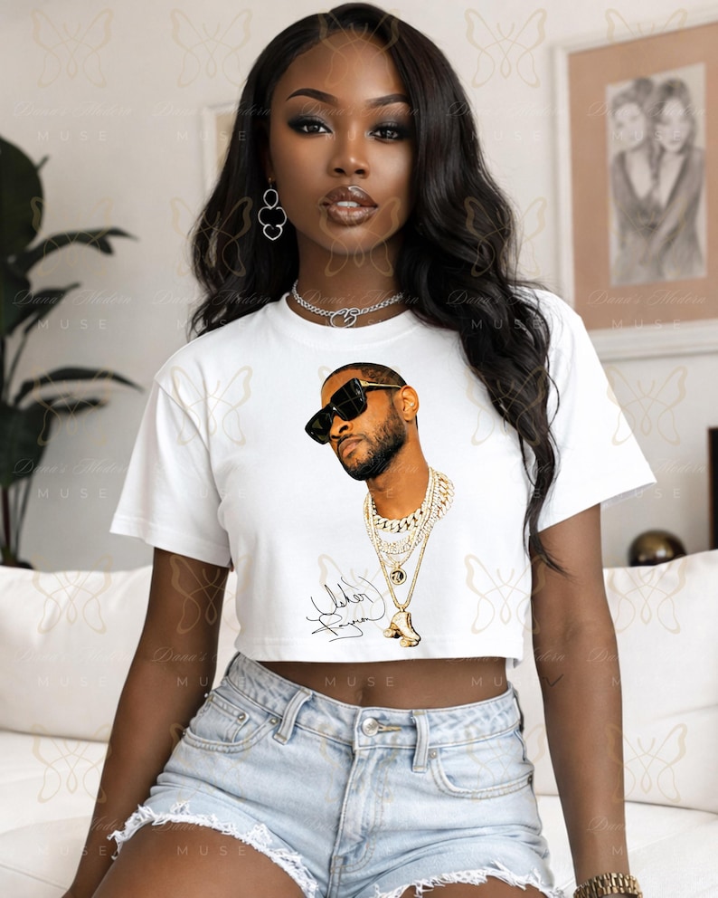 May include: A white short-sleeved crop top featuring a graphic print of a man wearing black sunglasses and multiple layered gold chains. Below the graphic is a black signature. The shirt has a crew neckline and a cropped hemline.