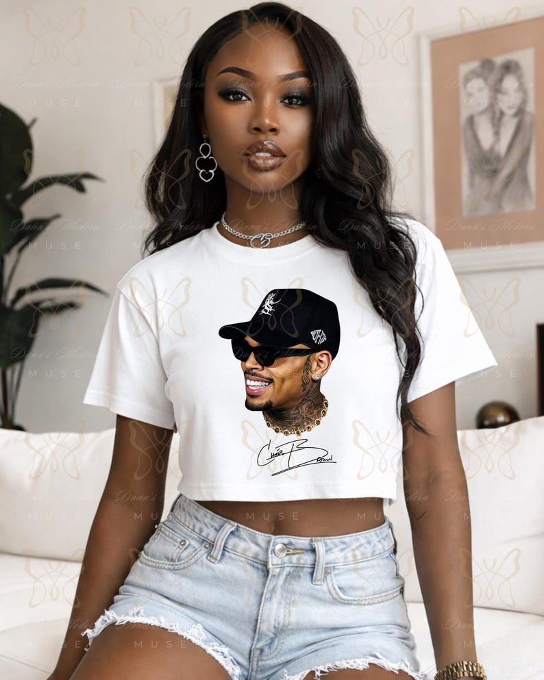May include: A white short-sleeved crop top featuring a graphic illustration of a person wearing a black baseball cap and dark sunglasses. The design includes a gold chain necklace and a signature script printed in black ink below the portrait.
