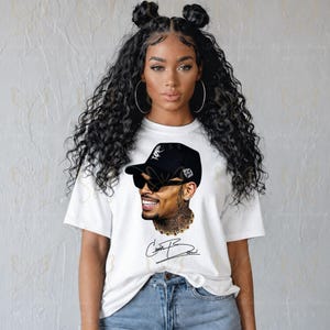 May include: A white short-sleeve t-shirt featuring a graphic print of a person wearing a black baseball cap and dark sunglasses. The print includes a gold chain necklace and the signature Chris Brown written in black script below the portrait.
