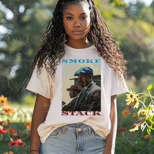 May include: A white t-shirt with a graphic of a vintage movie poster featuring two men in hats and the text "SMOKE STACK".