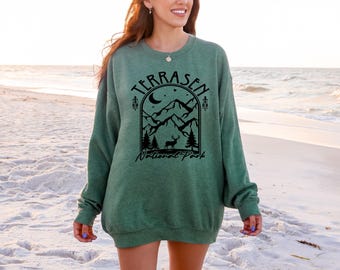 May include: Green sweatshirt with a black graphic of mountains, a deer, and the text "TERRASEN National Park." The long-sleeved top is made of a soft material and is suitable for casual wear.