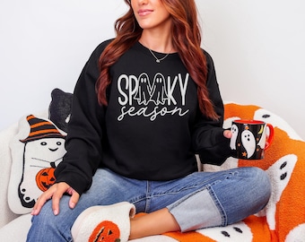 May include: Black sweatshirt with the words "Spooky Season" in white script and ghost graphics. A black mug with white ghost graphics and a red interior is held in the hand. The person is wearing blue jeans and white shoes.