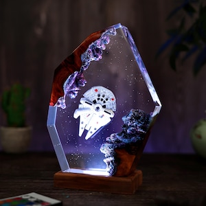 May include: A decorative resin lamp featuring the Millennium Falcon spaceship. The lamp has a clear, geometric shape with a wooden base and accents of brown, purple, and blue. The spaceship is white with red details, set against a starry background.