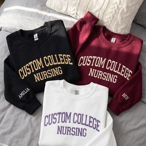 May include: Three sweatshirts in black, burgundy, and white, each with "CUSTOM COLLEGE NURSING" printed across the chest. The black sweatshirt has "AMELIA" on the sleeve, the burgundy has "BEN", and the white has "SARAH". The sweatshirts are on a gray bed.