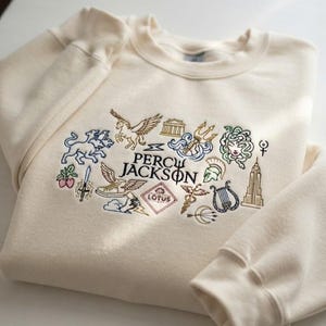 Peut inclure: Un sweat-shirt à col rond de couleur crème avec des icônes brodées colorées et le texte "PERCY JACKSON" au centre. La broderie comprend un chien à trois têtes, un cheval ailé, un trident, une lyre, un gratte-ciel et une fleur de lotus.