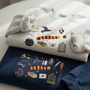 May include: A light grey sweatshirt and a navy blue sweatshirt, each featuring embroidered motifs including a wizard hat, a striped scarf, round glasses, a cauldron, a book, a letter with a red seal, a birdcage, a potion bottle, and a broom.