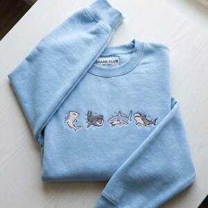 May include: Light blue sweatshirt with embroidered shark designs. The sweatshirt features four different shark illustrations in various colors. The text "SHARK CLUB EST. 2024" is printed on a small tag near the collar.