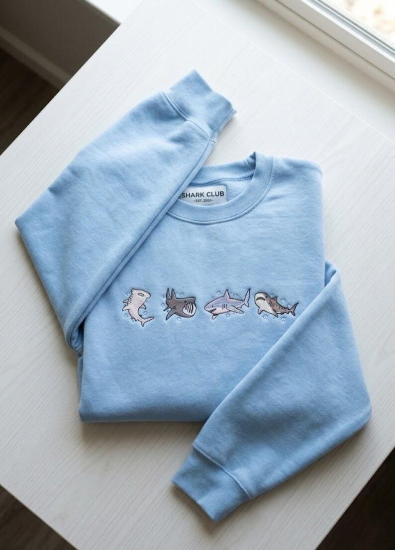 May include: Light blue sweatshirt with embroidered shark designs. The sweatshirt features four different shark illustrations in various colors. The text "SHARK CLUB EST. 2024" is printed on a small tag near the collar.