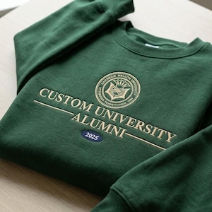 May include: A dark green sweatshirt with the text "CUSTOM UNIVERSITY ALUMNI" embroidered in a cream color. The sweatshirt also features a university emblem and the year "2025" in a blue oval. The garment is folded neatly on a light surface.