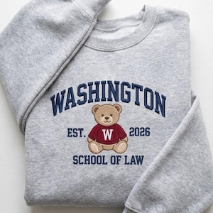 May include: A heather gray sweatshirt with the word "WASHINGTON" embroidered in navy blue. Below the word is a teddy bear wearing a red sweater with a white "W". The text "EST. 2026 SCHOOL OF LAW" is also embroidered on the sweatshirt.