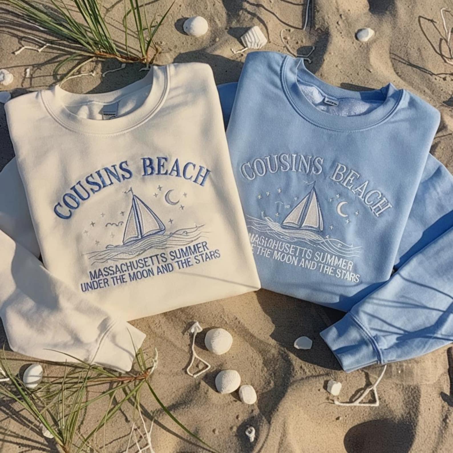 Two sweatshirts that read: Cousins Beach, Massachusetts Summer Under The Moon and The Stars. each sweatshirt also has an embroidered design of a sail boat sailing on waves with a moonlit, starry sky above it. The sweatshirt on the left is cream colored with its embroidered details in light blue and the one on the right is light blue featuring embroidered details in white. The sweatshirts are laid against a sandy background complete with seashells, grass, and twigs.