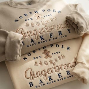 Peut inclure: Deux sweat-shirts beiges à col rond avec une broderie indiquant North Pole Est 1909 Gingerbread Bakery Hand Made With Love. Le motif comprend une icône de bonhomme en pain d'épices et de petites formes de Noël en fil bleu marine et marron.