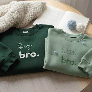 Könnte beinhalten: Ein dunkelwaldgrünes Sweatshirt mit dem weißen gestickten Text big bro. und ein helleres salbeigrünes Sweatshirt mit dem dunkelgrünen gestickten Text little bro. mit langen Ärmeln, gerippten Bündchen und einem Rundhalsausschnitt bei beiden.