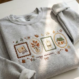 Könnte beinhalten: Ein hellgrauer Rundhals-Pullover mit gestickten Motiven eines offenen Buches mit Brille, einer Vase mit Herbstblättern, einer brennenden Kerze und einem kleinen Haus, jeweils in dekorativen Rahmen, mit dem Schriftzug "cozy season" darunter.