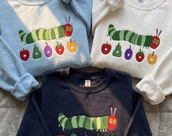 May include: Three crewneck sweatshirts in light blue, white, and navy blue, each featuring an embroidered green caterpillar with a red head and purple antennae, positioned above a row of five colorful embroidered fruits including an apple, pear, plum, strawberry, and orange.