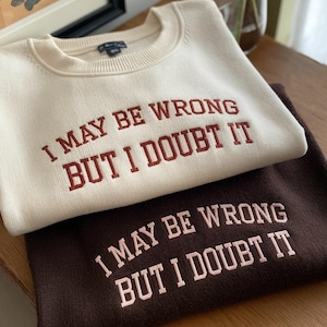 Puede incluir: Dos suéteres con el mismo texto, "I may be wrong but I doubt it", bordado en ellos. El suéter superior es de color crema con bordado marrón. El suéter inferior es marrón con bordado rosa.