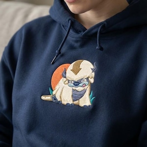 May include: A navy blue hooded sweatshirt featuring an embroidered graphic of a cream-colored, six-legged creature with a brown arrow marking on its head, a blue nose, and a tongue sticking out, set against an orange circular background.