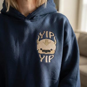 May include: A navy blue hooded sweatshirt featuring an embroidered design on the chest. The embroidery depicts a tan, horned creature with a brown arrow on its forehead, flanked by the text YIP above and YIP below in a light tan font.