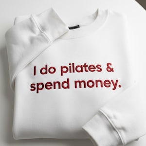 May include: A white crewneck sweatshirt featuring the embroidered red text "I do pilates & spend money." on the front. The garment has long sleeves with ribbed cuffs and a ribbed neckline, showcasing a clean, folded presentation of the fabric.