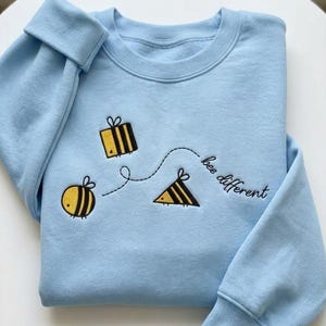 Op de afbeelding: Een lichtblauwe sweatshirt met geborduurde gele en zwarte bijen en de tekst "bee different". De sweatshirt heeft een ronde hals en lange mouwen met geribde manchetten. Het ontwerp toont drie gestileerde bijen die door een stippellijn met elkaar verbonden zijn.