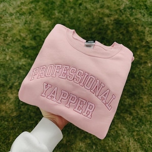 Puede incluir: Sudadera rosa con el texto "PROFESSIONAL YAPPER" bordado en una fuente de estilo universitario.