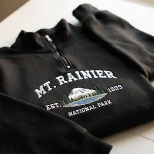 May include: A black quarter-zip sweatshirt featuring white embroidered text that reads MT. RAINIER EST. 1899 NATIONAL PARK. The center design includes an embroidered graphic of a snow-capped mountain, green pine trees, and a blue lake.