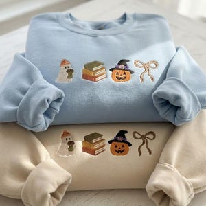 Può includere: Una felpa azzurra e una felpa beige, entrambe con un ricamo identico raffigurante un fantasma con berretto arancione, una pila di tre libri, una zucca con cappello da strega e un fiocco a nastro marrone centrato sul petto.