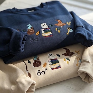 Puede incluir: Una sudadera azul marino y una sudadera beige, cada una con diseños bordados de un búho blanco sobre una pila de libros, un caldero marrón con vapor azul, un sombrero puntiagudo marrón, una llave dorada, una escoba y gafas redondas.