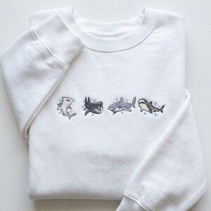 May include: White sweatshirt with embroidered shark designs. The crew neck sweatshirt features four different shark illustrations in shades of gray and blue. The sharks are embroidered across the chest of the folded garment.