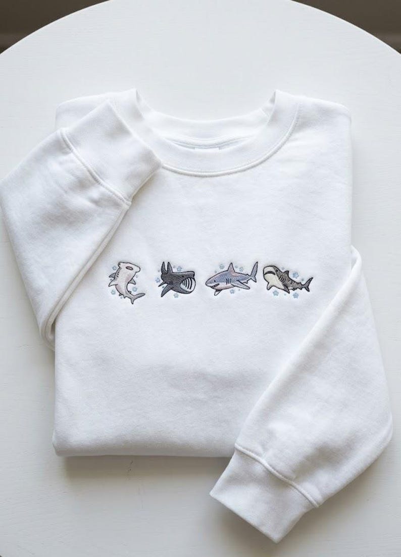 May include: White sweatshirt with embroidered shark designs. The crew neck sweatshirt features four different shark illustrations in shades of gray and blue. The sharks are embroidered across the chest of the folded garment.