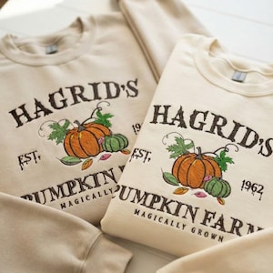 Może przedstawiać: Dwie beżowe bluzy z okrągłym dekoltem i ciemnobrązowym haftowanym napisem HAGRID'S PUMPKIN FARM EST. 1962 MAGICALLY GROWN. Wzór zawiera haftowaną ilustrację pomarańczowych i zielonych dyń z zielonymi liśćmi oraz małymi, kolorowymi listkami.