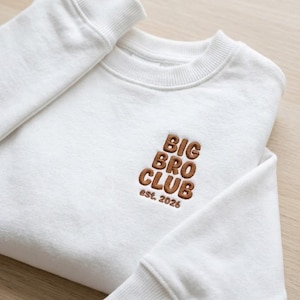 以下が含まれることがあります： 茶色の刺繍で「BIG BRO CLUB est. 2026」と書かれた白いクルーネックのスウェットシャツ。 スウェットシャツはリブ編みの袖口とクルーネックが特徴です。 生地は柔らかく快適に見えます。