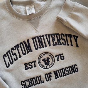 May include: Gray sweatshirt with black embroidered text that reads "Custom University Est 75 School of Nursing". The university logo is a black and white circle with a key and the letters "CBTT" around the outside.