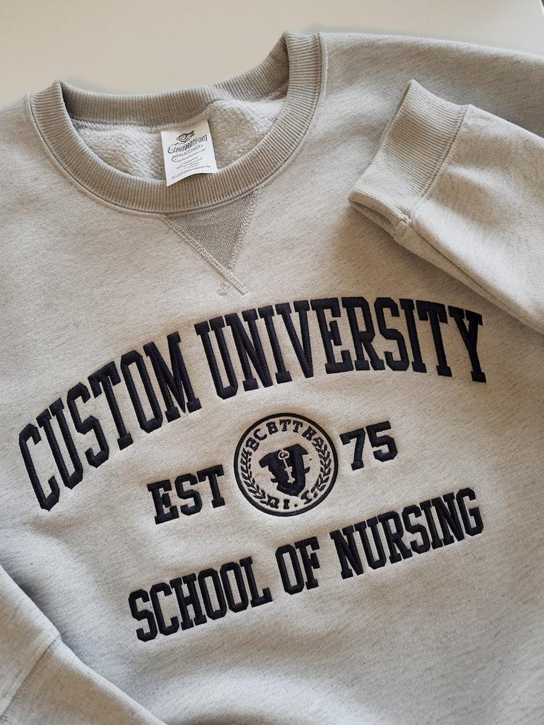 May include: Gray sweatshirt with black embroidered text that reads "Custom University Est 75 School of Nursing". The university logo is a black and white circle with a key and the letters "CBTT" around the outside.