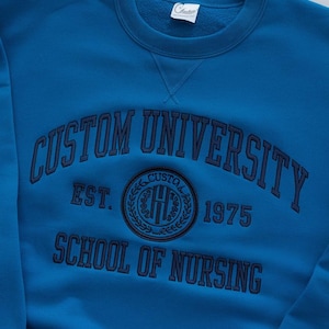May include: A blue sweatshirt with the text "Custom University Est. 1975 School of Nursing" in navy blue embroidery. A circular logo with the letters "CU" in a wreath is also embroidered on the sweatshirt.