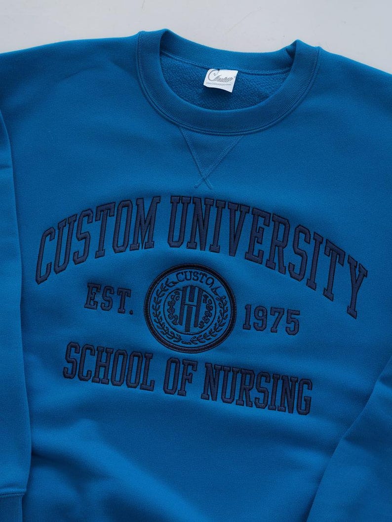May include: A blue sweatshirt with the text "Custom University Est. 1975 School of Nursing" in navy blue embroidery. A circular logo with the letters "CU" in a wreath is also embroidered on the sweatshirt.
