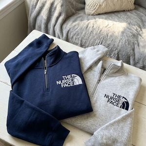 Op de afbeelding: Een marineblauwe en een grijs gemêleerde trui met kwartrits, elk voorzien van een wit of donkerblauw geborduurd logo op de linkerborst met de tekst THE NURSE FACE en een medisch caduceussymbool verwerkt in de bergafbeelding.