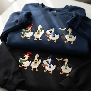 Puede incluir: Una sudadera azul marino y una sudadera negra, cada una con un diseño bordado de cuatro gansos blancos vestidos con atuendos de mago, incluyendo bufandas coloridas, sombreros y túnicas, con un pequeño caldero y una snitch dorada volando.