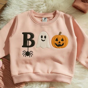 May include: A light pink sweatshirt with the word "BOO" in black and white embroidered letters and images. The "B" has a spider hanging from it. A white ghost and orange jack-o'-lantern are also embroidered on the front. A small brown beanie is in the background.