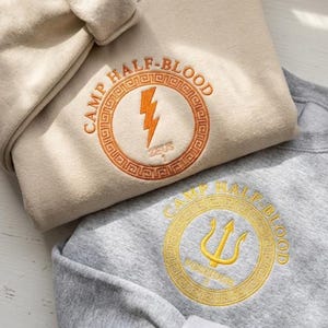 May include: A beige sweatshirt with orange embroidery featuring a lightning bolt and the text CAMP HALF-BLOOD ZEUS 1, and a grey sweatshirt with yellow embroidery featuring a trident and the text CAMP HALF-BLOOD POSEIDON 3, both with circular Greek key borders.