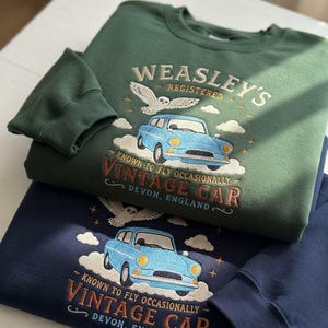 Op de afbeelding: Een bosgroen en een marineblauw sweatshirt, elk voorzien van een geborduurde lichtblauwe vintage auto, een witte uil en wolken. De tekst luidt: WEASLEY'S REGISTERED, KNOWN TO FLY OCCASIONALLY, VINTAGE CAR, DEVON, ENGLAND, met kleine gouden sterretjes.