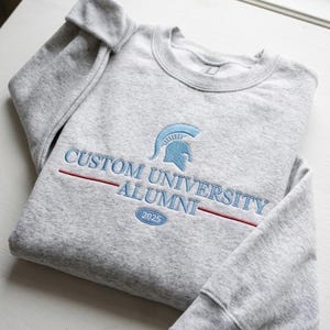 May include: A light gray crewneck sweatshirt featuring embroidered text that reads CUSTOM UNIVERSITY ALUMNI with a Spartan helmet icon above and the year 2025 inside an oval below, accented by a horizontal red line across the chest area.