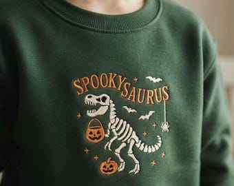 May include: A dark green crewneck sweatshirt featuring an embroidered design of a white dinosaur skeleton holding an orange jack-o'-lantern bucket. The text SPOOKYSAURUS is embroidered in orange above the dinosaur, surrounded by small bats, stars, and a spider.