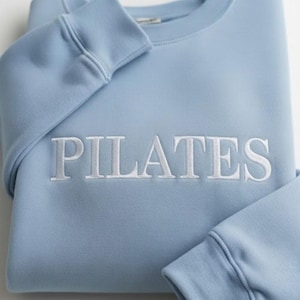May include: A light blue sweatshirt with the word "PILATES" embroidered in white across the chest. The sweatshirt has a crew neck and ribbed cuffs and hem. The sleeves are folded over.