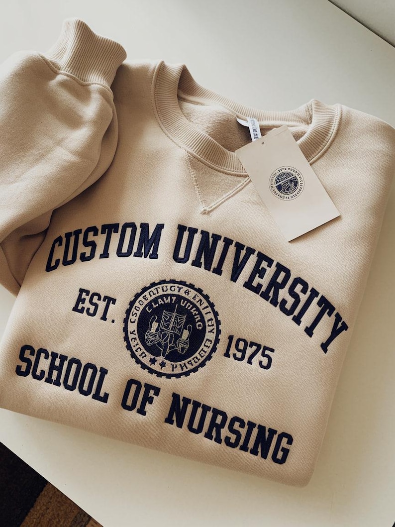May include: A beige sweatshirt with a navy blue embroidered design that reads "CUSTOM UNIVERSITY EST. 1975 SCHOOL OF NURSING". The design includes a circular emblem with a crest and the words "SOGEENTUGY & ENIHY LAWY U".