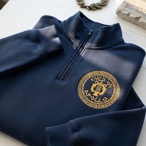 Może przedstawiać: Granatowa bluza z krótkim zamkiem błyskawicznym, ozdobiona na piersi złotym, okrągłym haftowanym emblematem. W centrum emblematu znajduje się lira, otoczona napisem "CHILD OF APOLLO" oraz wzorem greckiego meandra na zewnętrznej krawędzi.
