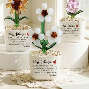 May include: Three decorative glass flower arrangements in white pots. One features a sunflower design, another has white flowers with brown centers, and the third has a pink flower. Each pot has text that reads "Mrs. Johnson" and a message.