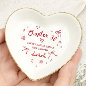 May include: A white heart-shaped trinket dish with a gold rim. The dish has red text that reads "Chapter 30" and "Every chapter brings new growth, Sarah". Decorative bows, flowers, and stars surround the text.
